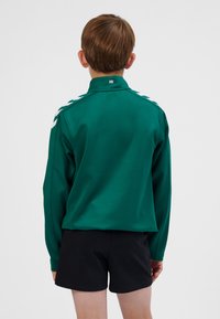 Grøn langærmet skjorte med høj krave, hvide skulderdetaljer og et logo ved halsen, kombineret med sorte shorts.