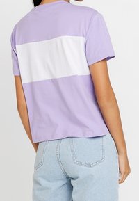 Short-sleeve lavender T-shirt with a white horizontal stripe across the back, made from soft cotton fabric, paired with light blue jeans.