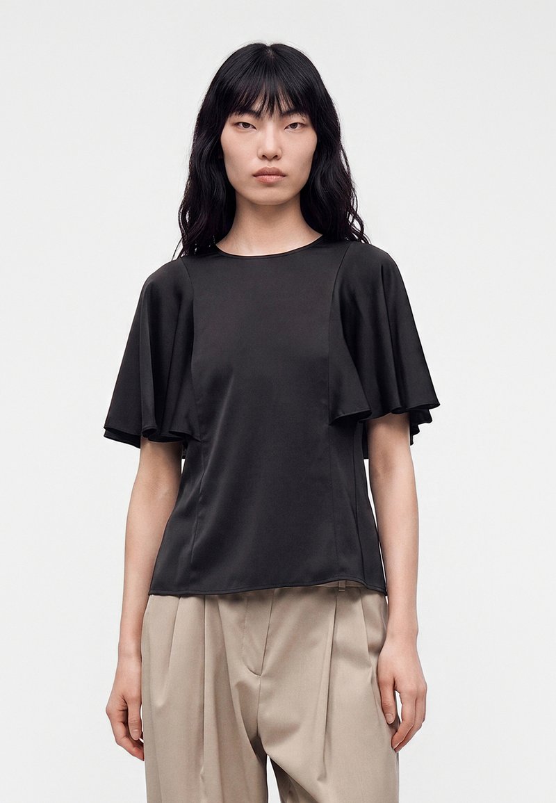 Femme aux longs cheveux noirs portant un haut noir à manches courtes avec manches volantes et un pantalon beige plissé taille haute, debout.