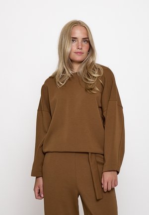 Giovane donna con lunghi capelli biondi che indossa una maglia marrone a maniche lunghe ampia e pantaloni abbinati, in piedi davanti a uno sfondo bianco semplice.