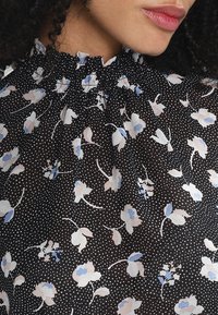 Czarna bluzka z marszczonym kołnierzykiem, z kwiecistym wzorem w jasnym różu i niebieskim oraz groszkami na półprzezroczystym materiale.