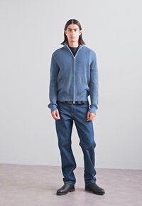 Μπλε ριγωτό φουτεράκι με φερμουάρ, μαύρο πουκάμισο, σκούρο μπλε τζιν, μαύρα καλτσάκια. Απλός σχεδιασμός, στενή σιλουέτα, ομαλή υφή. Απλό φόντο.