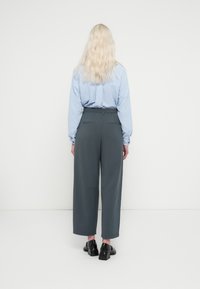 Femme aux cheveux longs blonds vue de dos, portant une chemise à manches longues bleu clair, un pantalon large taille haute gris foncé et des chaussures à talons noires.