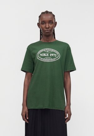 Punutiste juustega naine, kes kannab rohelist Nike 1972 t-särki ja musta seelikut, seisab ühevärvilise heleda tausta ees.