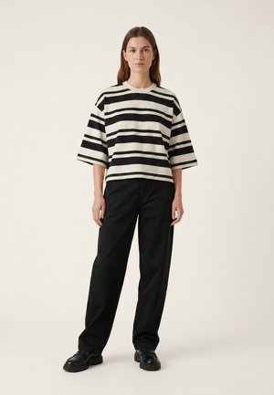 Nainen seisoo päällään mustavalkoinen leveäraita lyhythihainen paita, löysät mustat housut ja mustat massiiviset kengät yksinkertaisen vaalean taustan edessä.