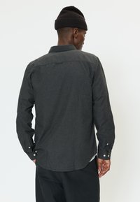 Hombre con camisa de manga larga abotonada gris oscuro y gorro negro, de pie con la espalda hacia la cámara.