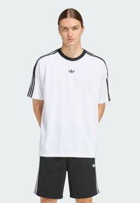 Balta T-kreka ar īsām piedurknēm, melna apkakle un tr�īs melnas svītras uz pleciem. Adidas logo uz krūtīm. Apvienota ar melniem īsiem šortiem.