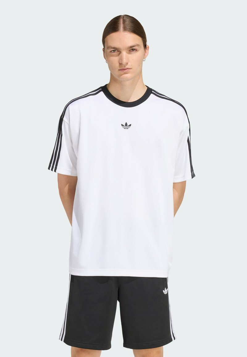 Camiseta blanca de manga corta con cuello negro y tres rayas negras en los hombros. Logo de Adidas en el pecho. Combinada con pantalones cortos negros.