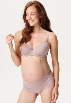 Glimlachende zwangere vrouw met lang haar die een bijpassende licht mauve zwangerschapsbeha en slipje draagt, staand tegen een effen achtergrond.