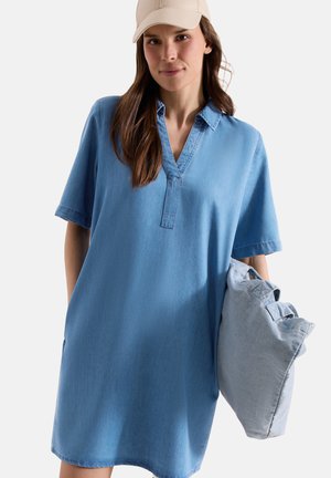 Femme portant une robe ample en denim bleu et une casquette beige, tenant un sac cabas en denim bleu clair sur fond blanc.