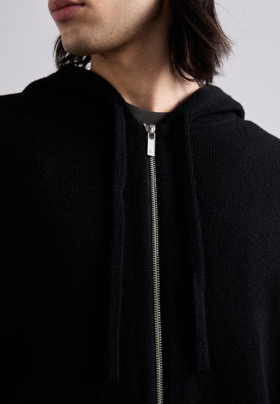 Schwarze Zip-Hoodie aus einem weichen, strukturierten Material. Verfügt über einen silbernen Reißverschluss und Zugbänder, mit Kapuze und gerippten Kanten.