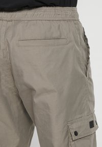 Beige cargo hlače s sproščenim krojem, elastičnim pasom, stranskimi žepi in ravnim zadnjim žepom. Imajo črne kovinske dodatke.