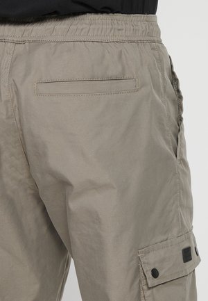 Beige cargo nadrág laza fit, elasztikus derékkal, oldalsó zsebekkel és lapos hátsó zsebbel. Fekete hardver részletek jellemzik.
