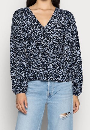 Blusa nera con scollo a V, caratterizzata da un motivo a goccia blu e bianco, maniche lunghe a sbuffo e vita arricciata. Indossata con jeans azzurri.