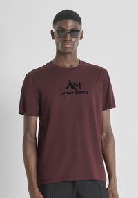 Joven con gafas de sol de montura transparente, una camiseta burdeos de Antony Morato y pantalones negros, de pie contra un fondo claro y sencillo.