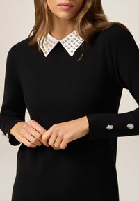 Schwarzes Strickkleid mit einem weißen, besetzten Kragen und silbernen Knopfakzenten an den Ärmeln, das ein schlankes und strukturiertes Design betont.