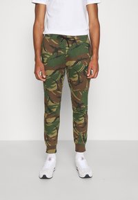 Kamouflage-joggers i grönt och brunt tyg med resårmidja, dragsko och ribbade mudd, tillsammans med vita sneakers.