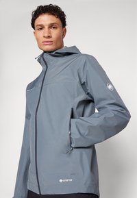 Grau GORE-TEX-Jacke mit Kapuze, Reißverschluss, Seitentaschen und dezentem Logo-Detail. Glatter Stoff und wasserdichtes Design.