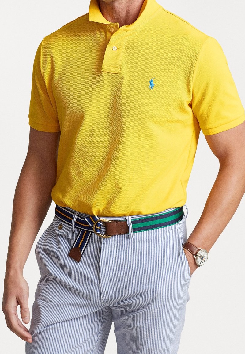 Homme portant un polo jaune à manches courtes, un pantalon à rayures blanches et bleues, une ceinture rayée bleu-vert, et une montre-bracelet argentée.