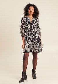 Schwarz-weißes Blumendruckkleid mit drei Viertel-Ärmeln, einem gefältelten Ausschnitt und einem ausgestellten Saum. Kombiniert mit schwarzen Stiefeletten.