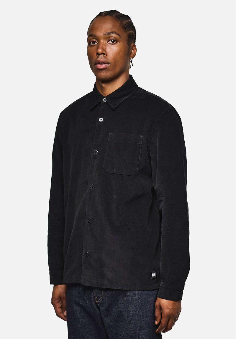Camicia a bottone in corduroy nero con maniche lunghe, taschino sul petto e una trama sottile. Presenta bottoni bianchi e un piccolo etichetta del marchio sull'orlo.