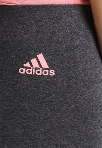 Szare legginsy sportowe wykonane z miękkiego materiału, z różowym logo Adidas po lewej stronie u dołu. Gładka faktura i dopasowany krój.