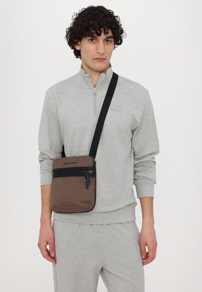 Sac bandoulière zippé marron avec motif de logo embossé, zips noirs et sangle ajustable, porté sur un sweat à zip gris clair.