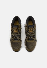 Zapatillas deportivas de color verde oliva oscuro y negro con una parte superior texturizada, punta redondeada, acentos amarillos en contraste y cordones con un patrón.
