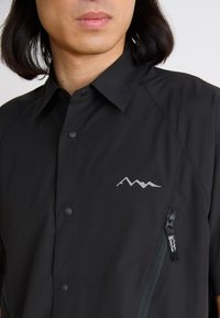 Black button-up shirt featuring a subtle mountain logo in silver, zippered pocket on the side, made from a smooth, lightweight material.