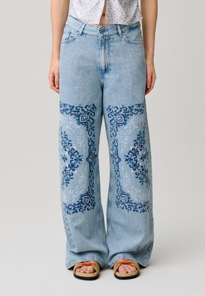 Jean large bleu clair avec broderie géométrique bleu foncé sur les cuisses avant, porté avec des sandales camel et une chemise courte à fleurs.