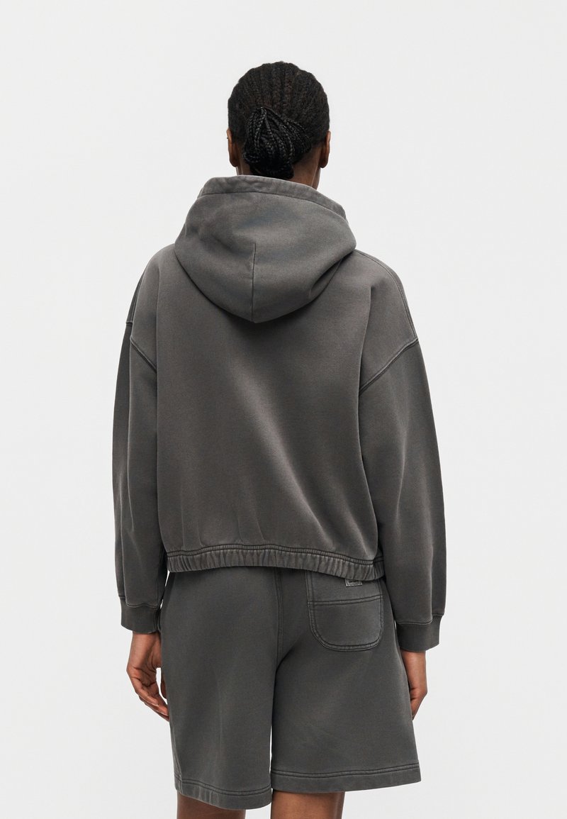 Persona con i capelli intrecciati che indossa una felpa con cappuccio grigio scuro e pantaloncini abbinati, vista da dietro su uno sfondo uniforme.