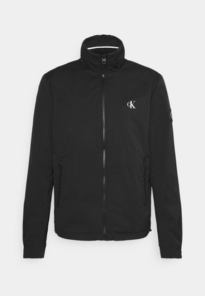 Veste noire zippée avec col montant, poches latérales, logo Calvin Klein sur la poitrine et petite écusson sur la manche gauche, présentée à plat sur fond blanc.