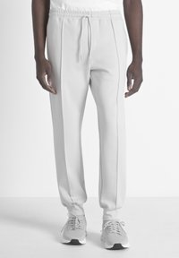 Pantalons de survêtement gris clair à taille élastique avec cordon, présentant des coutures latérales et une coupe décontractée. Associés à des chaussures de sport gris clair.