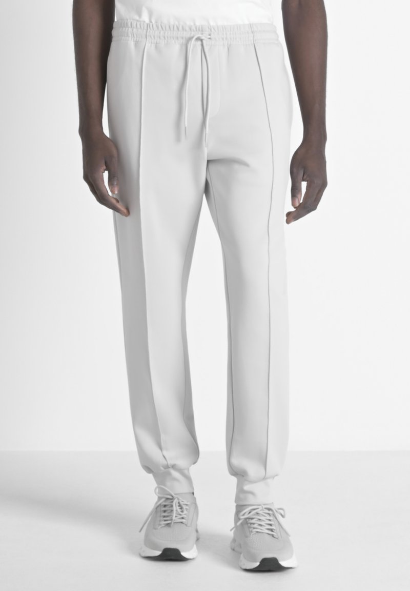 Pantalons de survêtement gris clair à taille élastique avec cordon, présentant des coutures latérales et une coupe décontractée. Associés à des chaussures de sport gris clair.