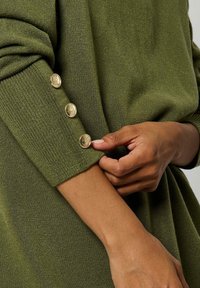 Maglione a maglia verde oliva con maniche a coste e accenti di bottoni dorati sui polsini, che mette in risalto un tessuto testurizzato e un design aderente.