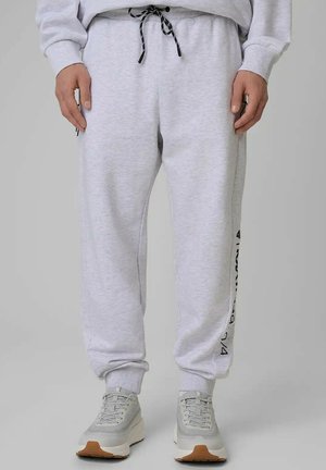 Osoba nosząca jasnoszare spodnie dresowe z czarnym sznurkiem i bocznym napisem, połączone z szarymi trampkami z białymi podeszwami i brązowymi podeszwami zewnętrznymi.