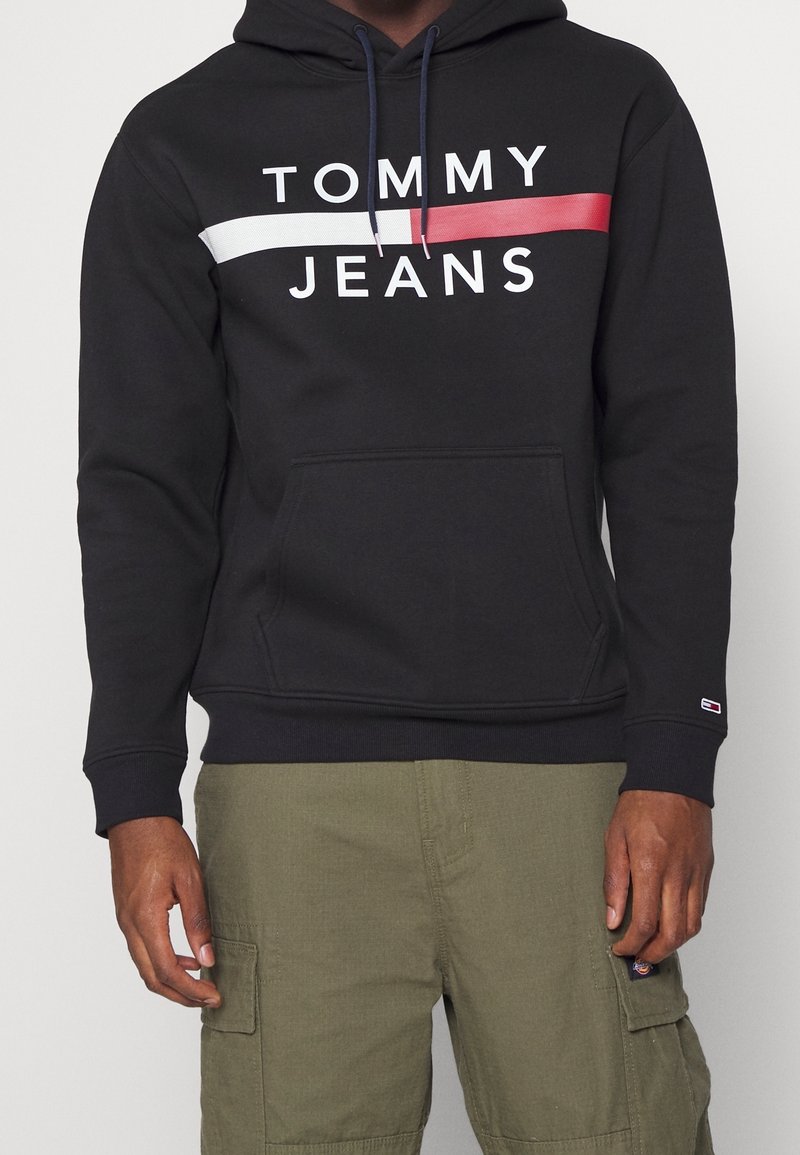 Černá mikina s klokaní kapsou, s bílými, červenými a modrými vodorovnými pruhy a nápisem "TOMMY JEANS" v tučném písmu na hrudi.