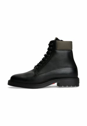 Bota de tobillo negra de cuero con cordones, con un cuello acolchado gris oscuro y suela gruesa que presenta un detalle rojo, blanco y azul cerca del talón.