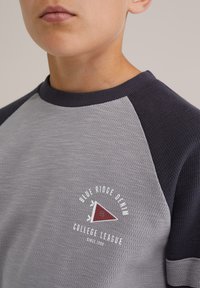 Szürke és sötétkék raglán póló, texturált anyagból. Háromszög alakú logóval, alatta "Blue Ridge Denim College League" felirattal.