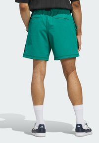 Groene shorts met een elastische tailleband, twee achterzakken, contrasterende stiksels en een logo-patch aan de zijkant. Draagt met witte sokken en sneakers.