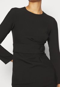 Femme portant une robe noire moulante à manches longues avec une large ceinture à la taille, ornée d'une grande boucle ronde recouverte de tissu.
