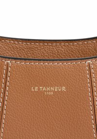 Materiale in pelle di un caldo marrone con finitura testurizzata. Presenta cuciture bianche e un logo in rilievo dorato con la scritta "LE TANNEUR 1898."