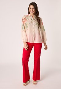 Bloemdruk blouse met knopen en puff mouwen in lichtroze, met rode en groene botanische prints. Gecombineerd met rode flared broek en beige hakken.