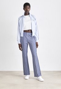 Light blue track jacket with black accents, paired with patterned blue flared pants. Model wears white sneakers and a cropped white top.