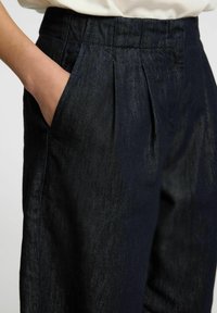 Un pantalon en denim foncé présente une coupe ajustée, un devant plat, un pli visible et des poches latérales. Le tissu a un léger éclat et une texture lisse.