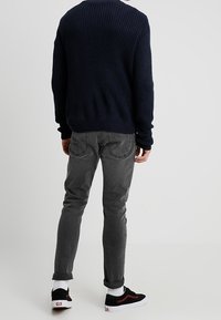 Marineblauer Strickpullover mit gerippter Textur, kombiniert mit schmalen grauen Jeans. Schwarze Sneakers mit roten Akzenten und weißen Sohlen. Lässiges Outfit, Rückansicht.