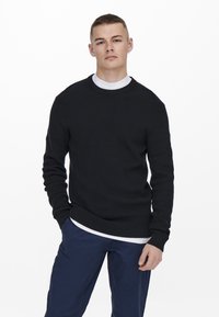 Pull noir côtelé avec un col rond, associé à une chemise blanche en dessous. Le mannequin porte un pantalon bleu marine, les mains dans les poches.