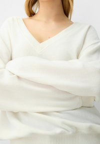 Biały, oversize'owy sweter z dekoltem w serek, prążkowaną fakturą i długimi rękawami, podkreślający luźny krój oraz miękki materiał.