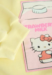 Tela amarilla con puño acanalado y un parche rosa que muestra un gato de dibujos animados sosteniendo un cartón de leche de fresa con el texto "STRAWBERRY MILK".