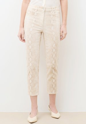 Kobieta ubrana w beżowe spodnie o prostych nogawkach w wężowy wzór i kremowe buty na kwadratowym nosku, stojąca na tle jednolitego białego tła.